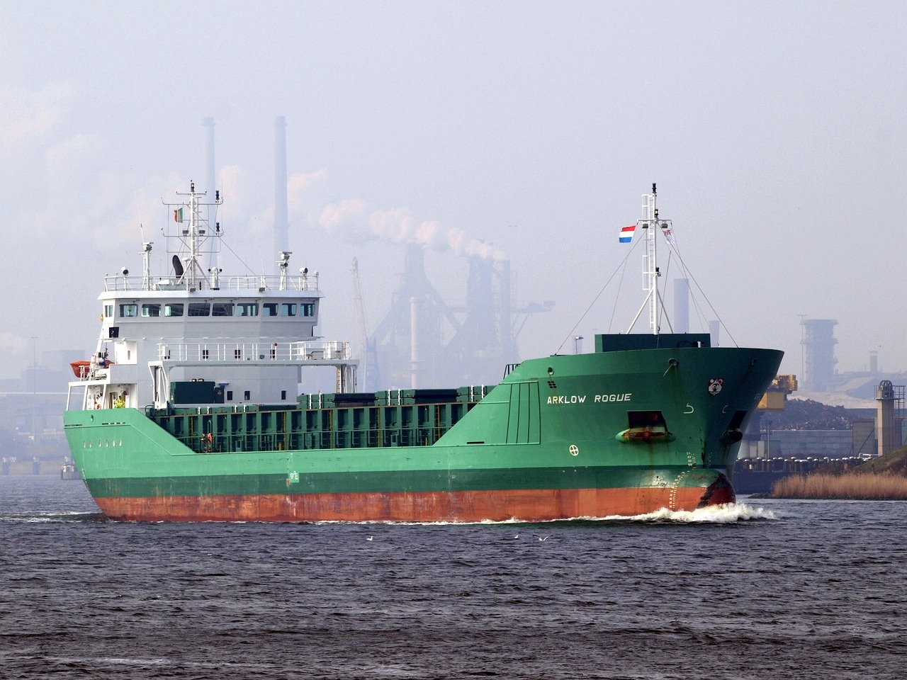 Stückgutschiff vor industrieller Hafenkulisse mit Schornsteinen und Kaianlagen