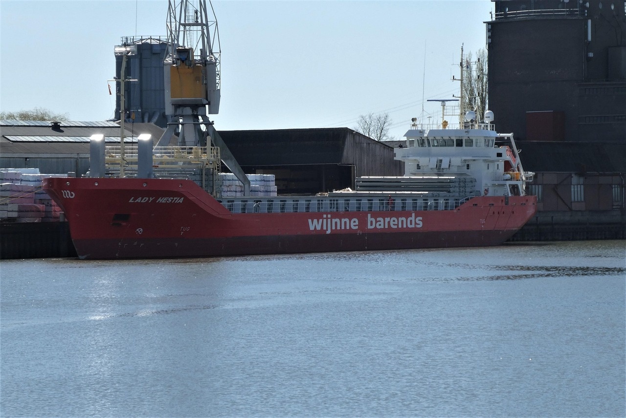 Stückgutschiff am Kai vor Hafenkran und industriellen Lagerhallen